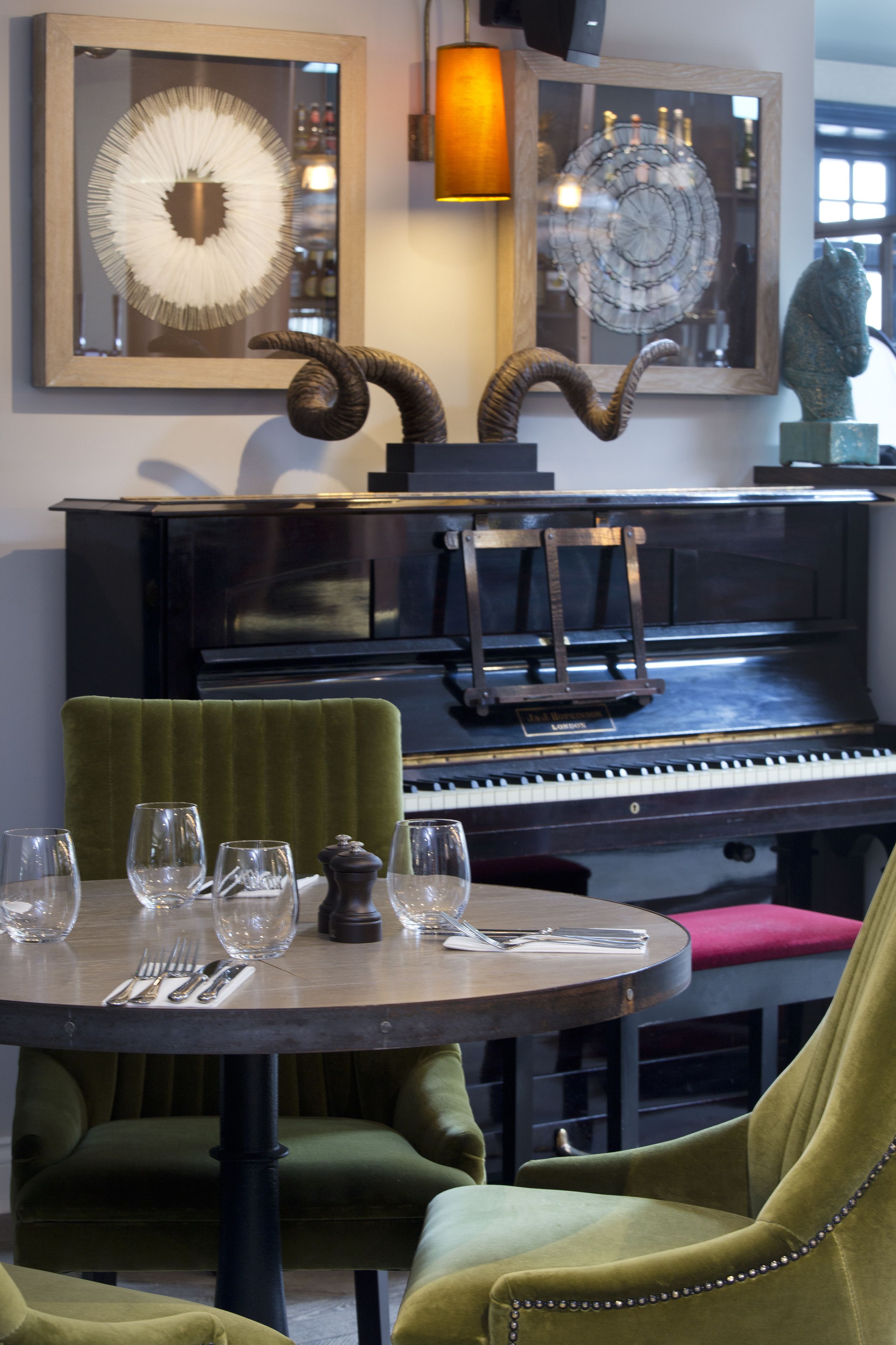 A dining area with a piano, green upholstered chairs, a round table set with wine glasses and cutlery, and decorative items including a spiral sculpture and framed artwork.