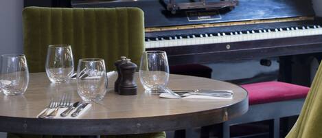 A dining area with a piano, green upholstered chairs, a round table set with wine glasses and cutlery, and decorative items including a spiral sculpture and framed artwork.