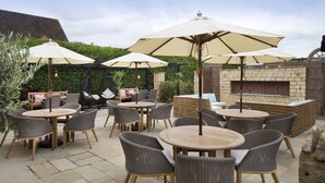 Eine Terrasse im Freien mit runden Tischen und Korbstühlen, beschattet durch große Sonnenschirme, und einem Kamin aus Stein.