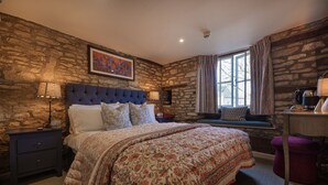 Ein Schlafzimmer mit Steinstruktur, einem gepolsterten Kopfteil, einem großen Bett mit floralem Bettbezug, einem Nachttisch, einem Fenster mit Vorhängen und einem Schreibtisch mit Lampe.