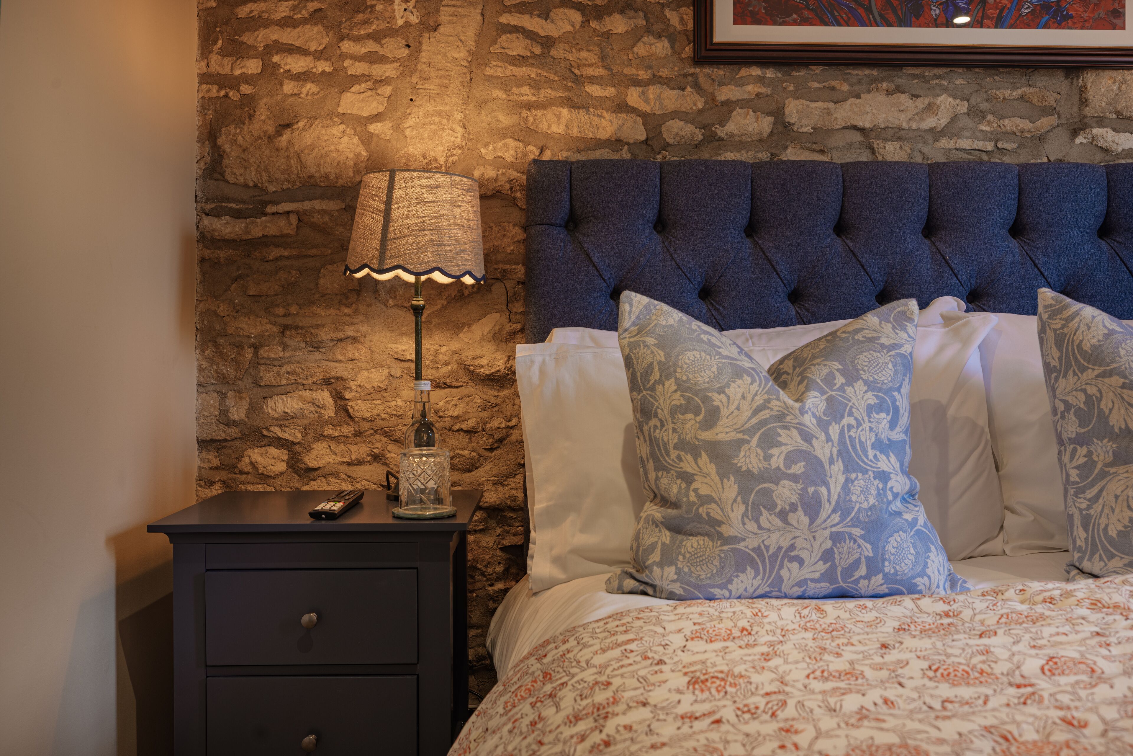 A bedroom with a stone wall, a dark nightstand with a lamp, and a bed with patterned pillows.