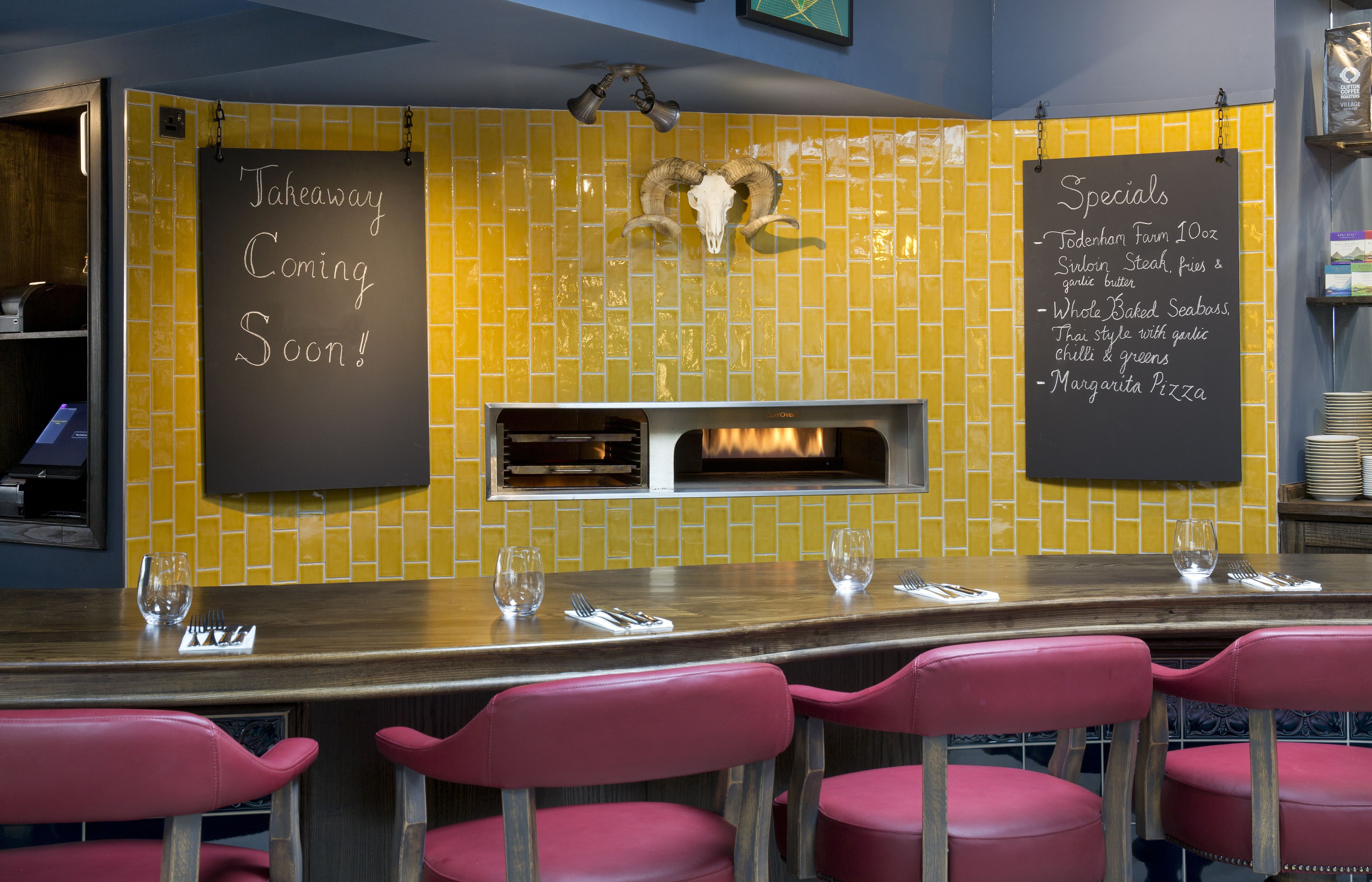 A restaurant with a yellow-tiled wall, a blackboard with specials, and a pizza oven.