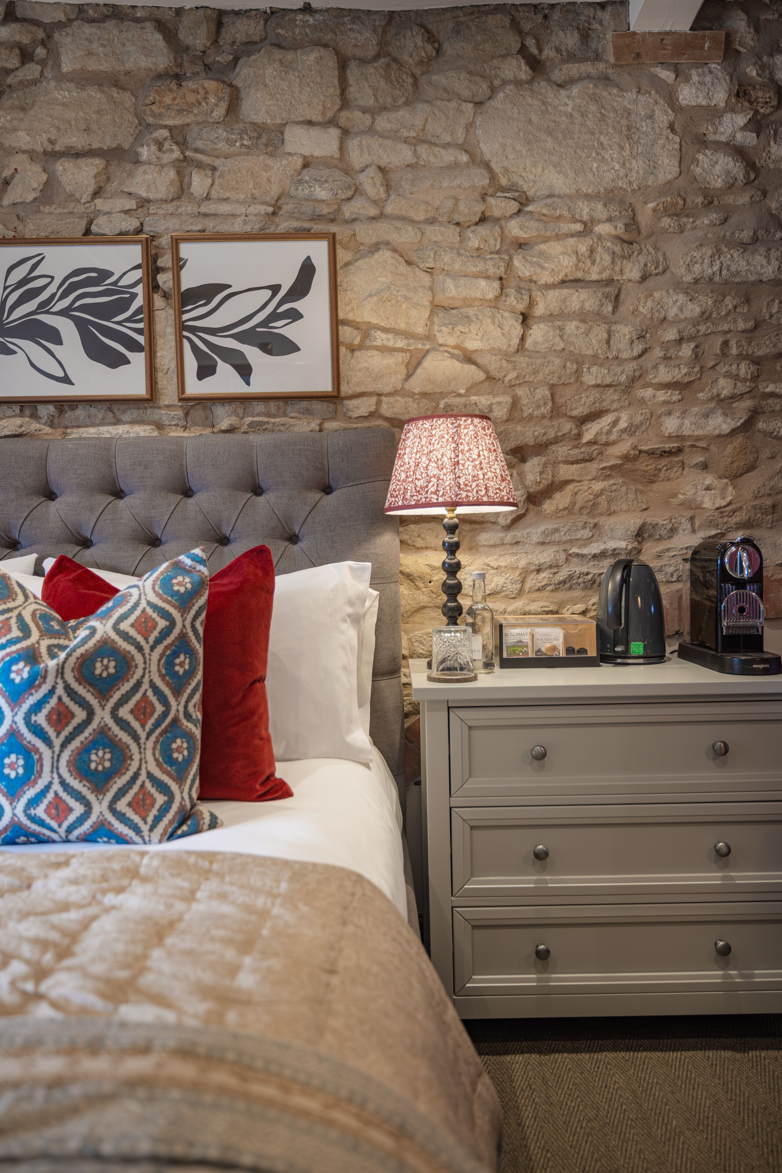 A bedroom with a stone wall, a tufted headboard, a bed with patterned pillows, a bedside table with a lamp, a coffee maker, and framed artwork.