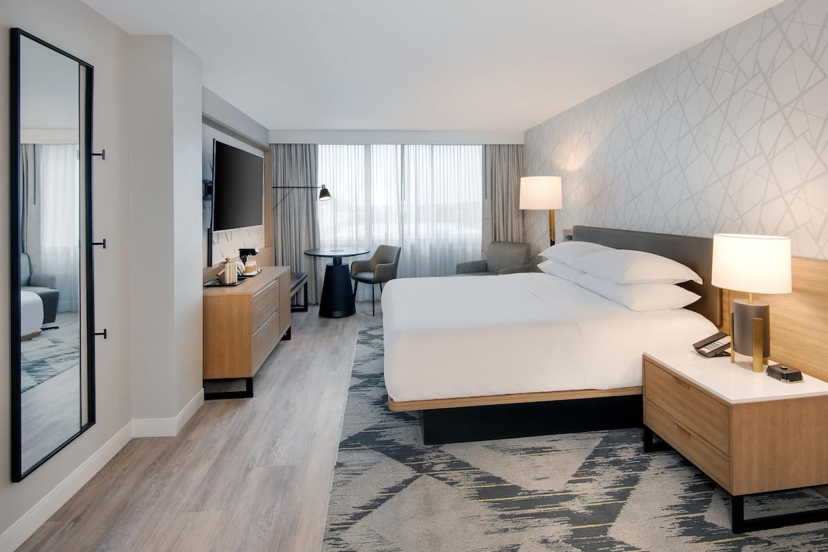 A hotel room with a large bed, bedside tables, a desk with a chair, a TV, and a view of the outdoors.