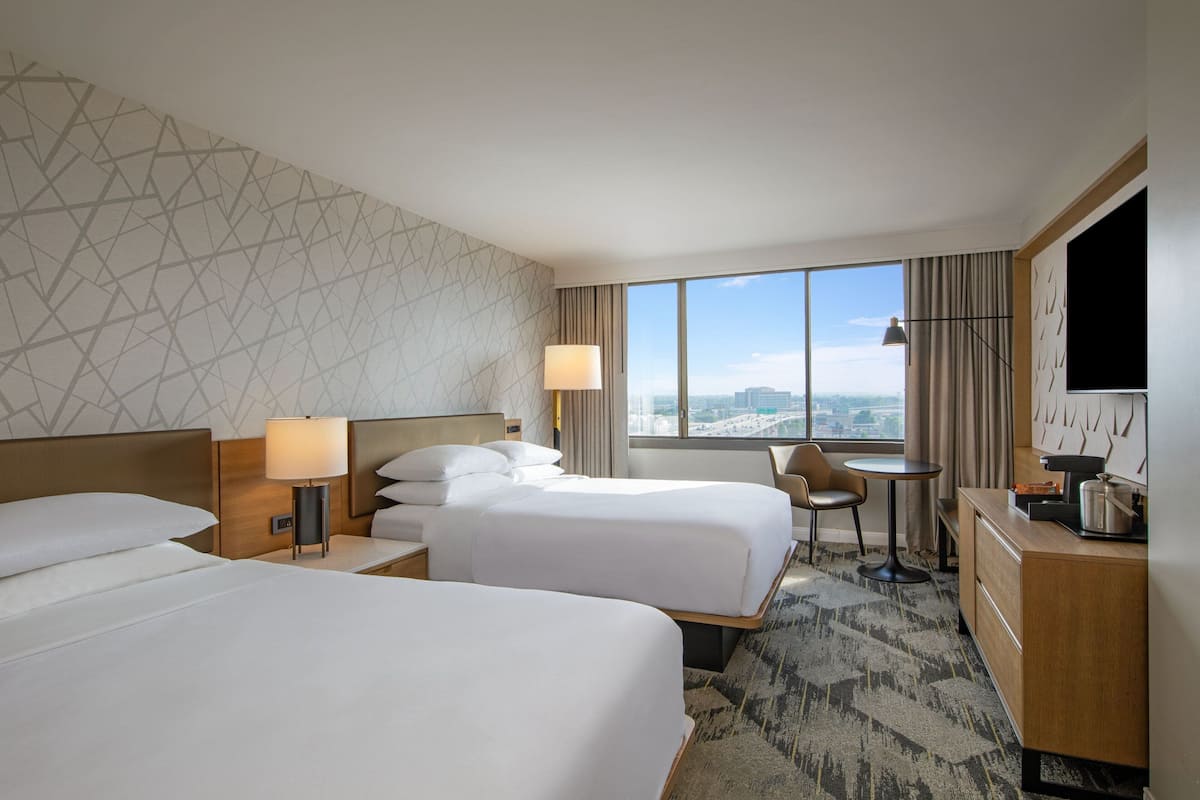 A hotel room with two beds, a desk, a chair, a TV, and a large window with a city view.