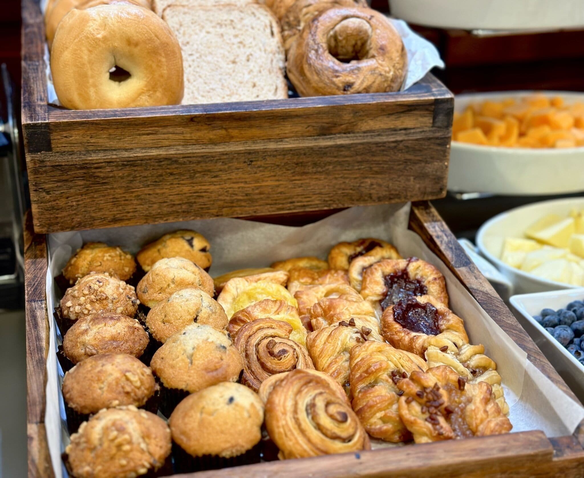 A wooden serving tray with various baked goods including bagels, bread, muffins, and pastries.