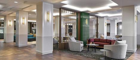 A spacious lobby with a modern design, featuring a wooden herringbone floor, a seating area with sofas and armchairs, and a decorative partition with vertical lines.