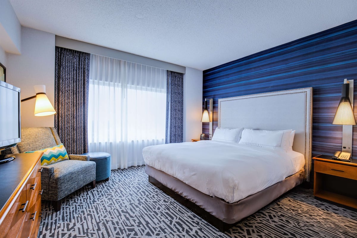 A hotel room with a large bed, a desk with a lamp, a chair, and a patterned carpet.