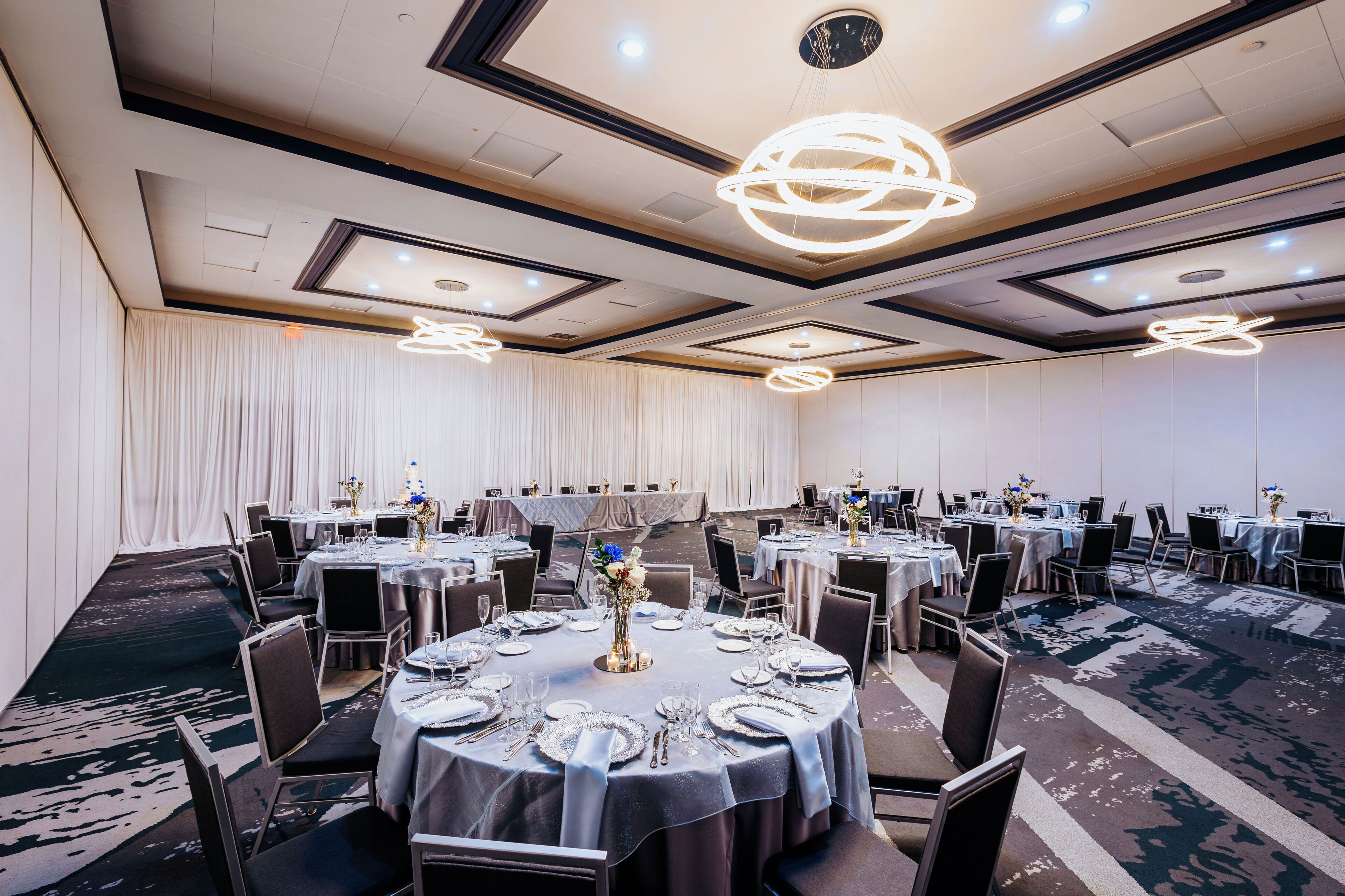 A banquet hall with round tables set for dining, featuring a modern chandelier and a carpeted floor.