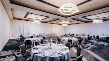 A banquet hall with round tables set for dining, featuring a modern chandelier and a carpeted floor.