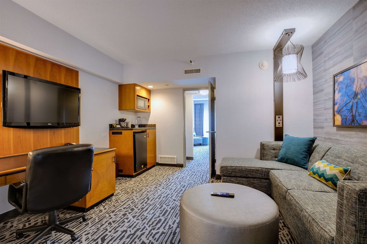 A hotel room with a flat-screen TV mounted on the wall, a kitchenette with a microwave and refrigerator, a gray sofa with a patterned throw pillow, and a round ottoman with a remote control on it.