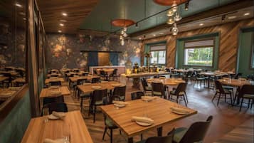 A restaurant with wooden tables and chairs, a bar area, and large windows with a view of greenery.