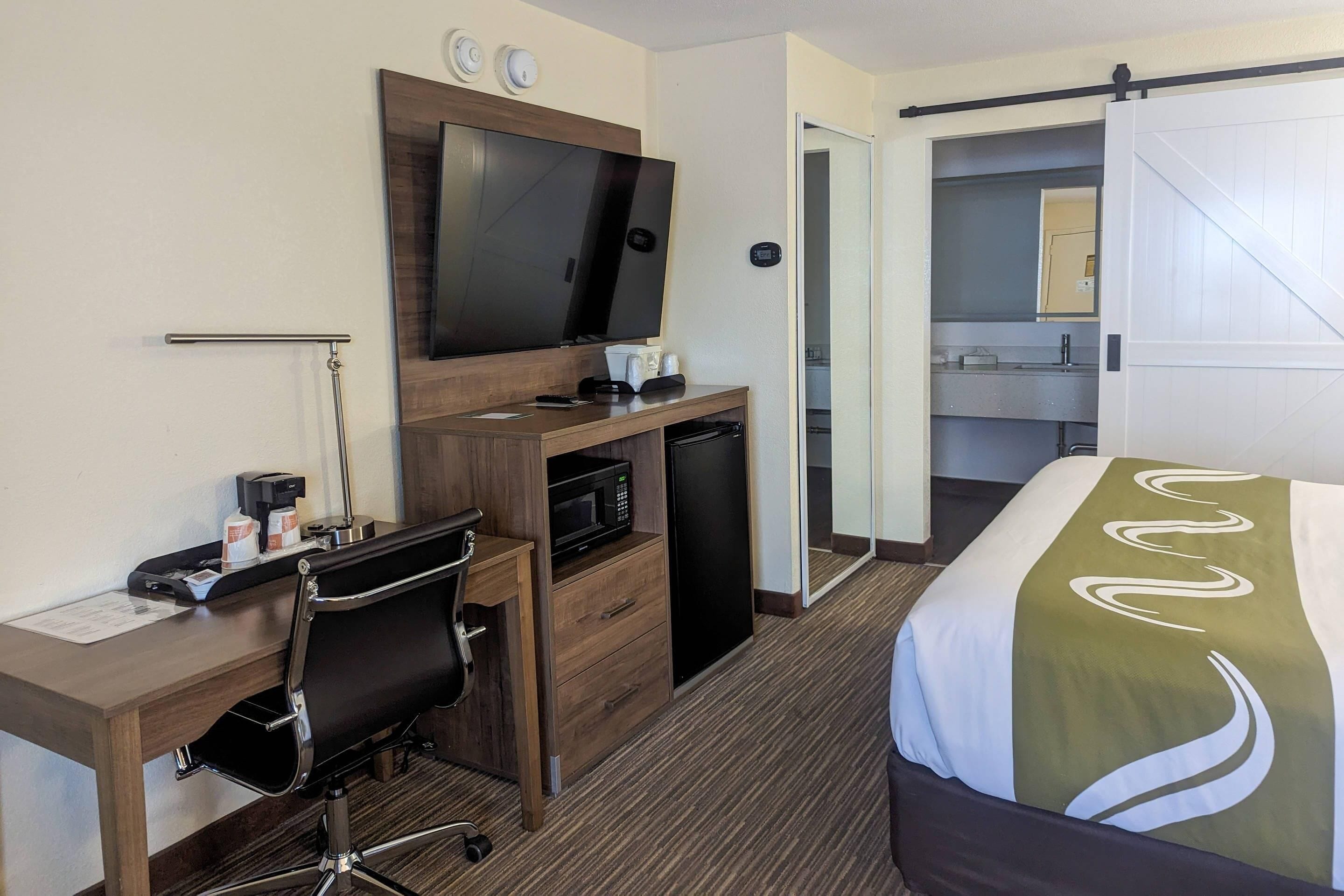 Ett hotellrum med en säng, ett skrivbord med en stol, en tv, en mikrovågsugn och ett litet kylskåp.
