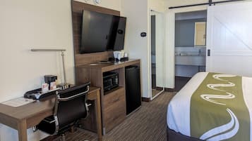 Ett hotellrum med en säng, ett skrivbord med en stol, en tv, en mikrovågsugn och ett litet kylskåp.