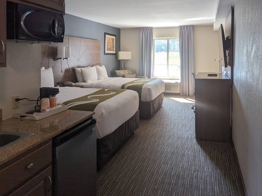 A hotel room with two beds, a microwave, a small table with a phone and a coffee maker, a sink, and a large window with curtains.