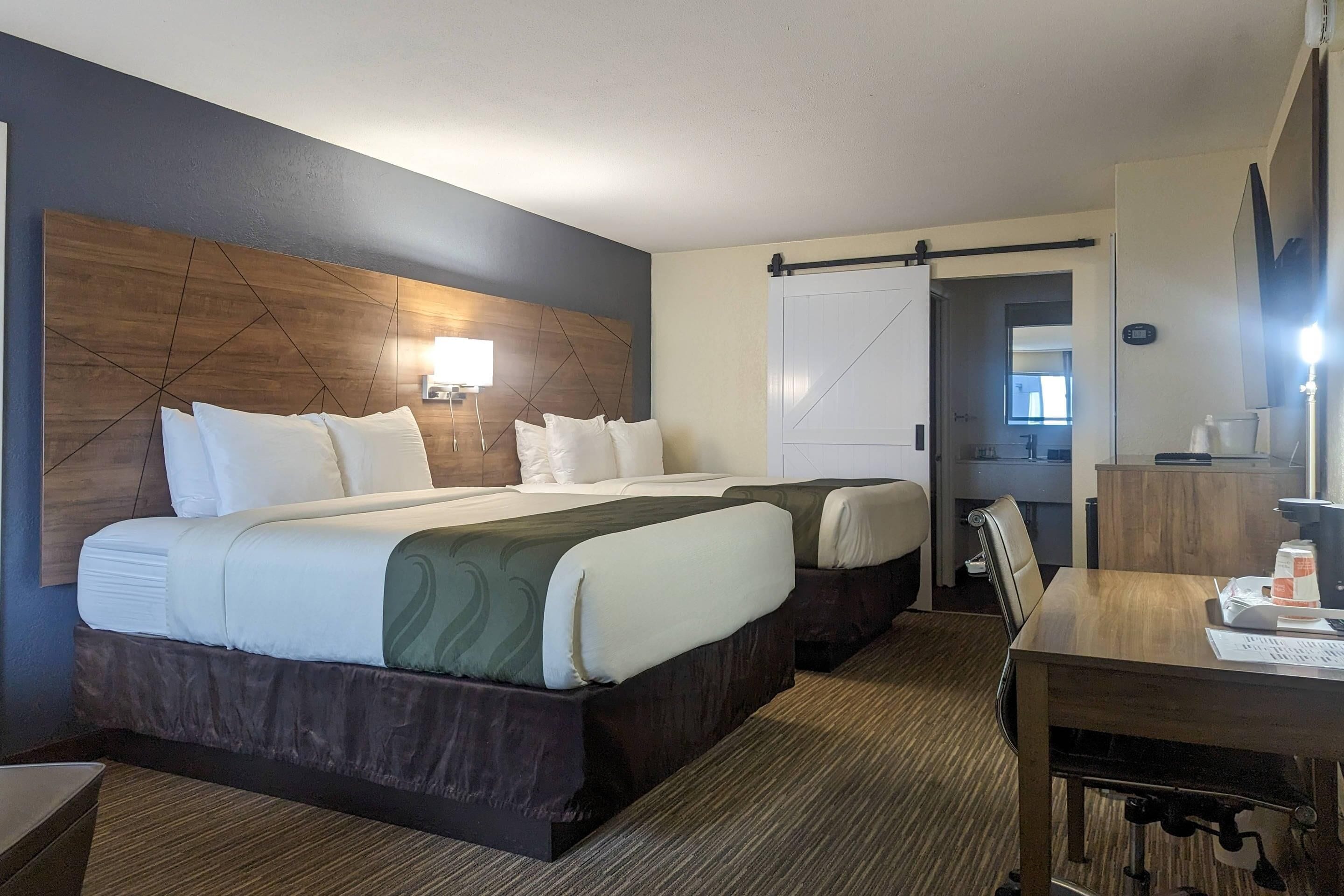 Ein Hotelzimmer mit zwei Betten, einem Schreibtisch, einem Stuhl, einer Lampe und einer Schiebetür zu einem Badezimmer.