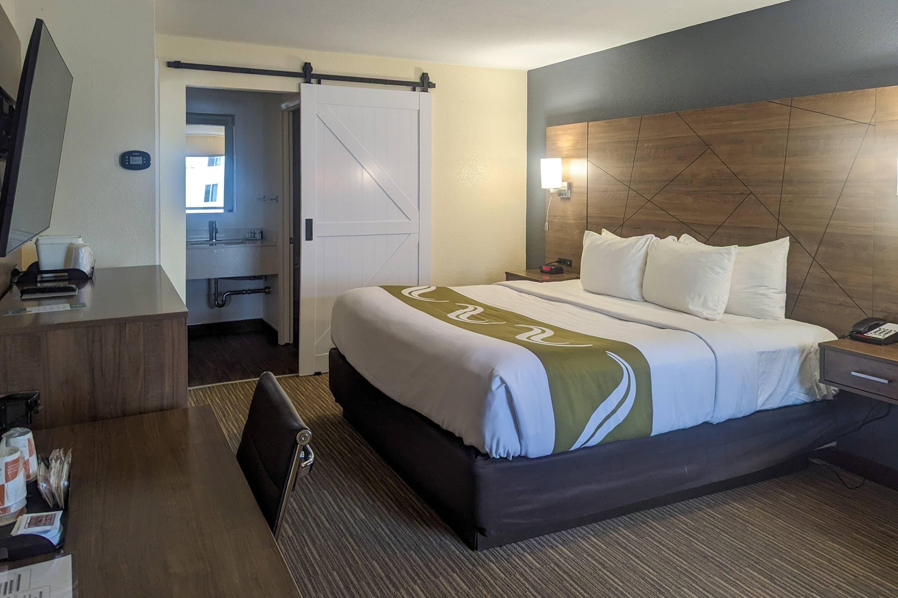 A hotel room with a bed, bedside table, desk, chair, and a bathroom visible through an open door.