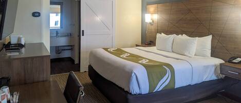 A hotel room with a bed, bedside table, desk, chair, and a bathroom visible through an open door.