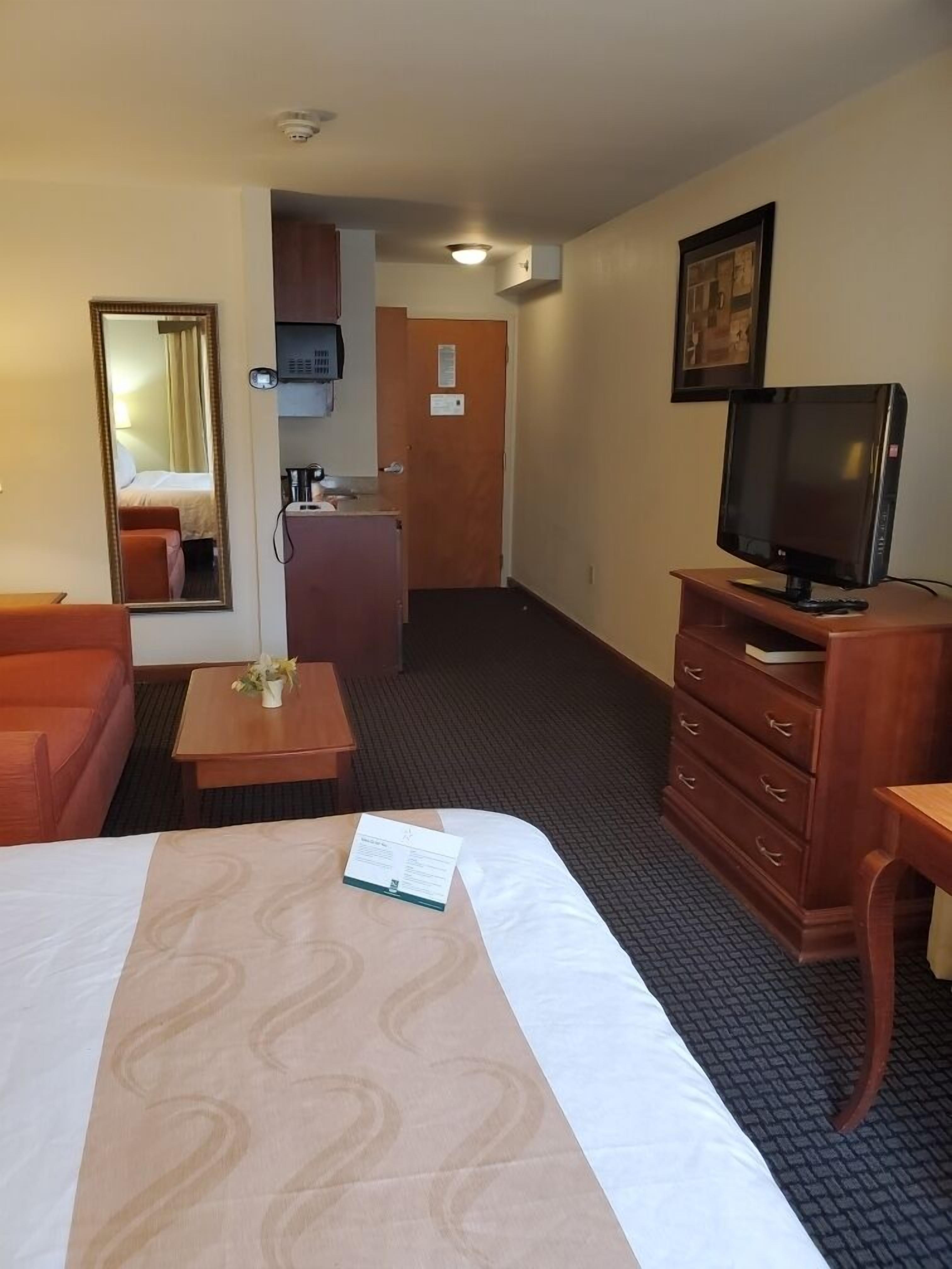 A hotel room with a bed, a desk, a television, a mirror, and a kitchenette.