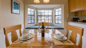 A dining area with a wooden table set for four, a bottle of wine, and a view through large windows. - Channel Island Shores (Oxnard)