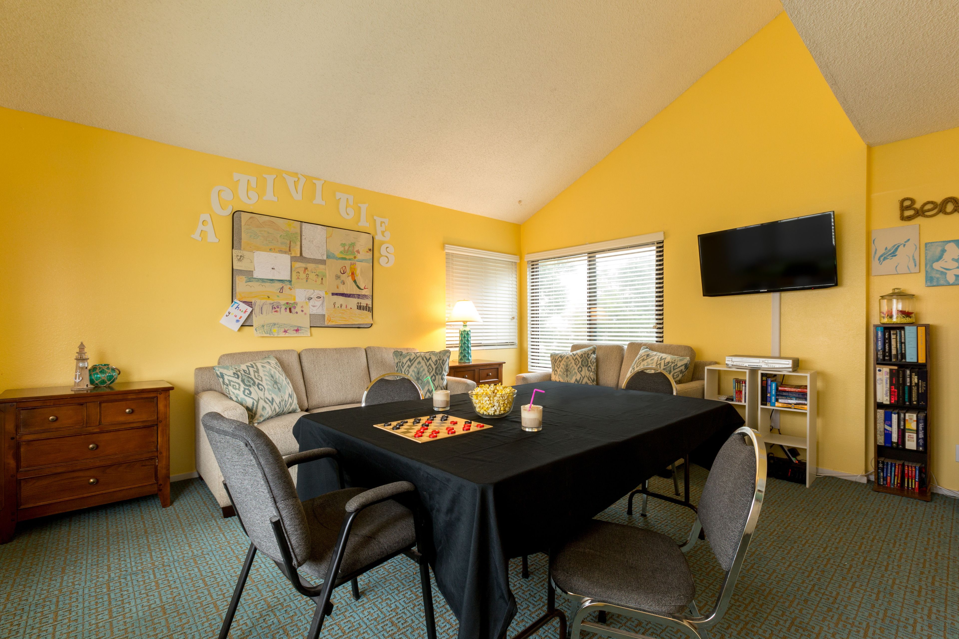 A room with a yellow wall, a bulletin board, a television, a bookshelf, and a dining table with chairs.