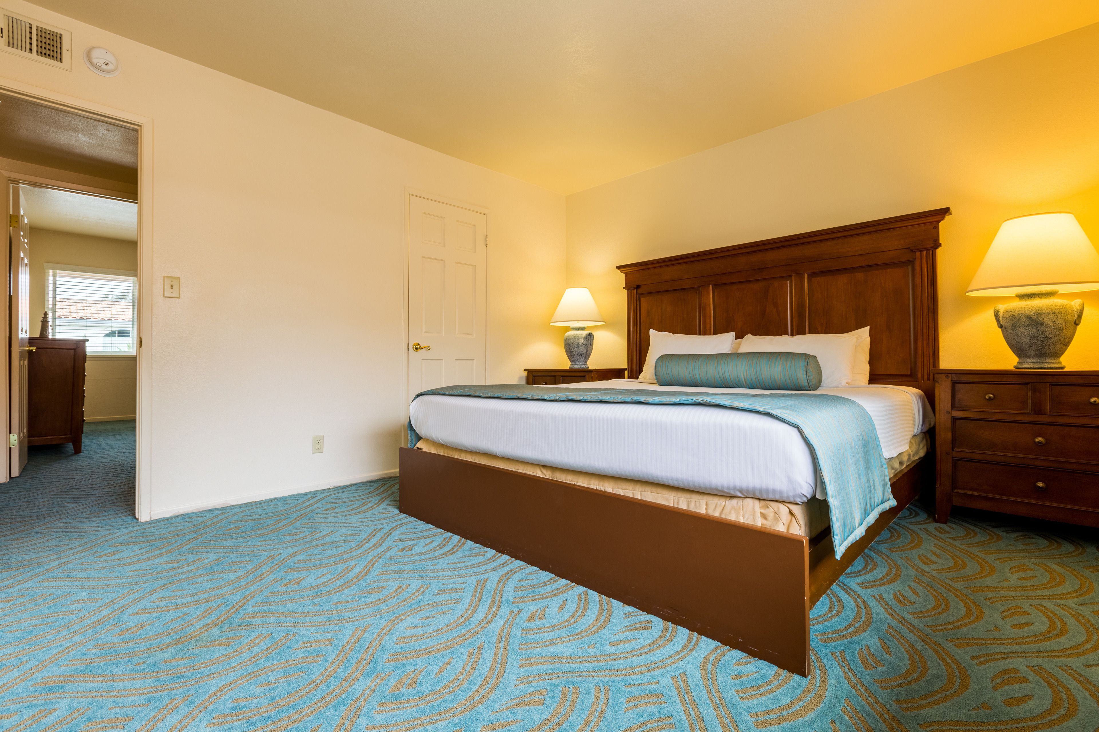 Habitación de hotel con una cama grande, cabecera de madera, dos mesitas de noche con lámparas, una puerta que conduce a otra habitación y una alfombra estampada.