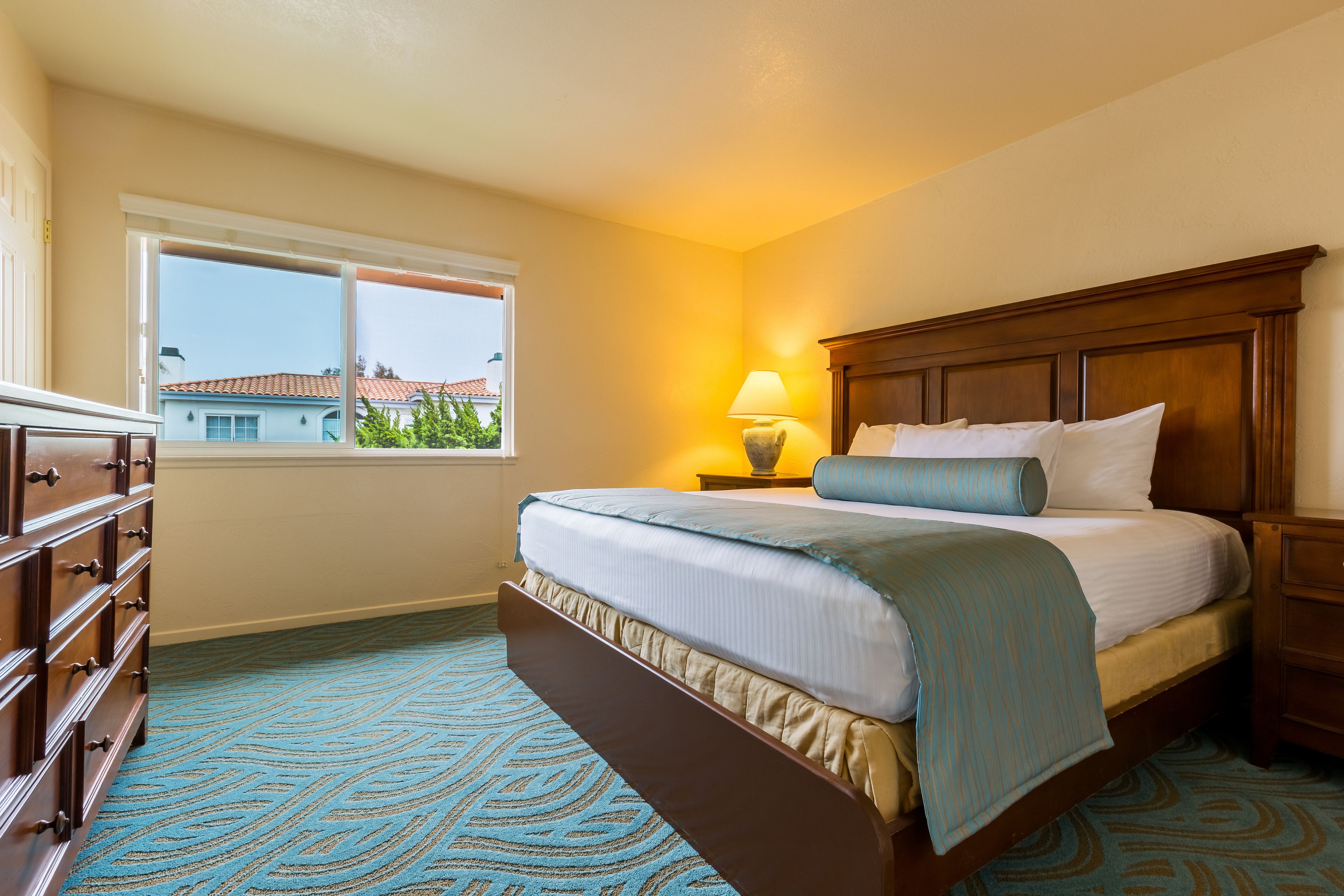 A bedroom with a bed, dresser, nightstand, lamp, and window with a view of buildings.