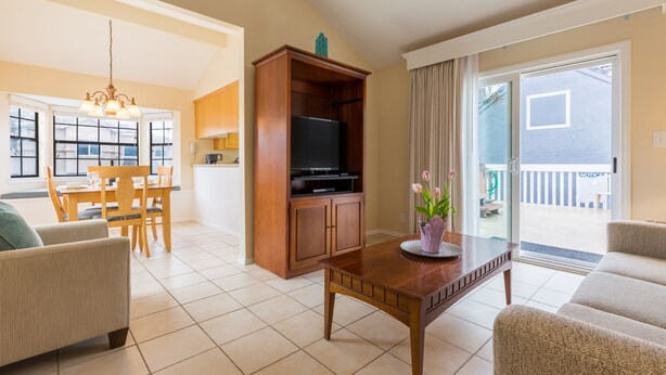 A living room with a wooden TV stand, a dining table with chairs, a sofa, and a sliding glass door leading to a balcony. - Channel Island Shores (Oxnard)