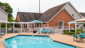 A swimming pool with lounge chairs and umbrellas, surrounded by a fenced area with houses in the background.