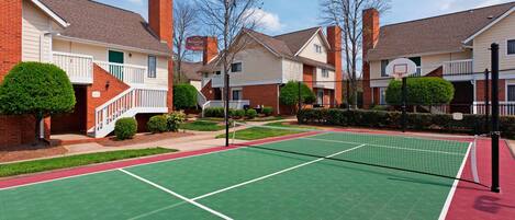 Una cancha de tenis con red, rodeada de edificios residenciales con fachadas de ladrillo y césped verde.
