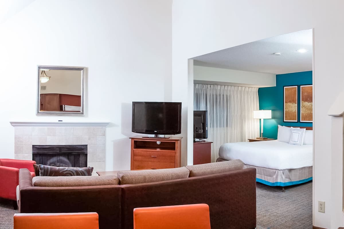 A hotel room with a living area featuring a sofa, a flat-screen TV, and a fireplace. The room has a bed with a blue accent wall and a staircase.