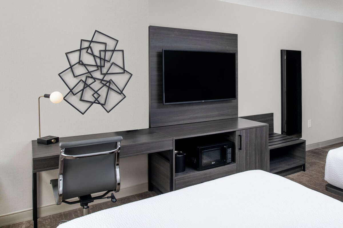 Une chambre d’hôtel moderne dotée d’un téléviseur à écran plat fixé sur un panneau en bois foncé, d’une œuvre d’art murale géométrique noire, d’un bureau avec une chaise et d’un micro-ondes encastré.