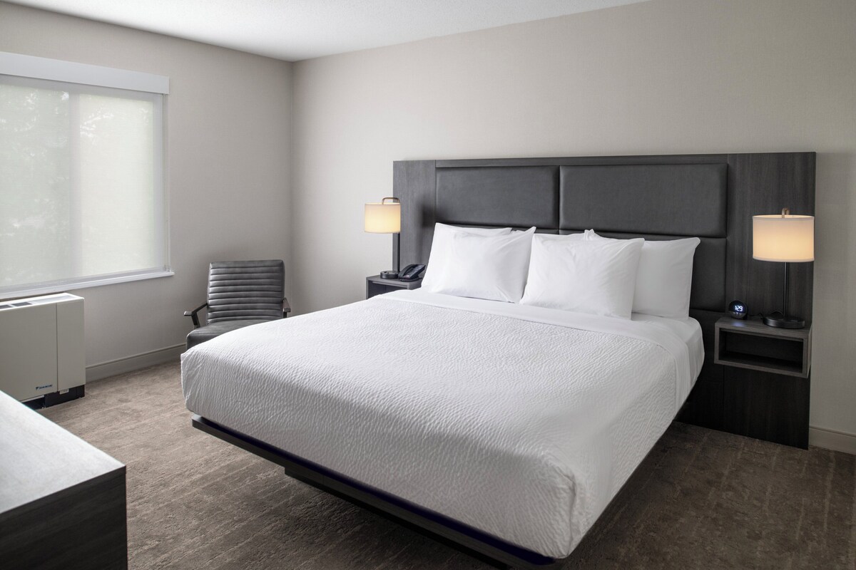Une chambre d’hôtel moderne avec un grand lit, deux tables de chevet, une chaise et une fenêtre avec des stores.