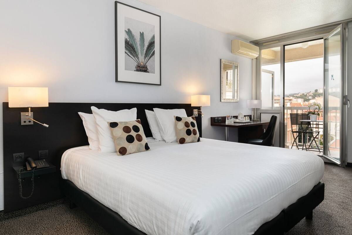 Ein Hotelzimmer mit einem großen Bett, zwei Nachttischlampen, einem Schreibtisch mit Stuhl, einem großen Fenster und einem gerahmten Bild an der Wand.