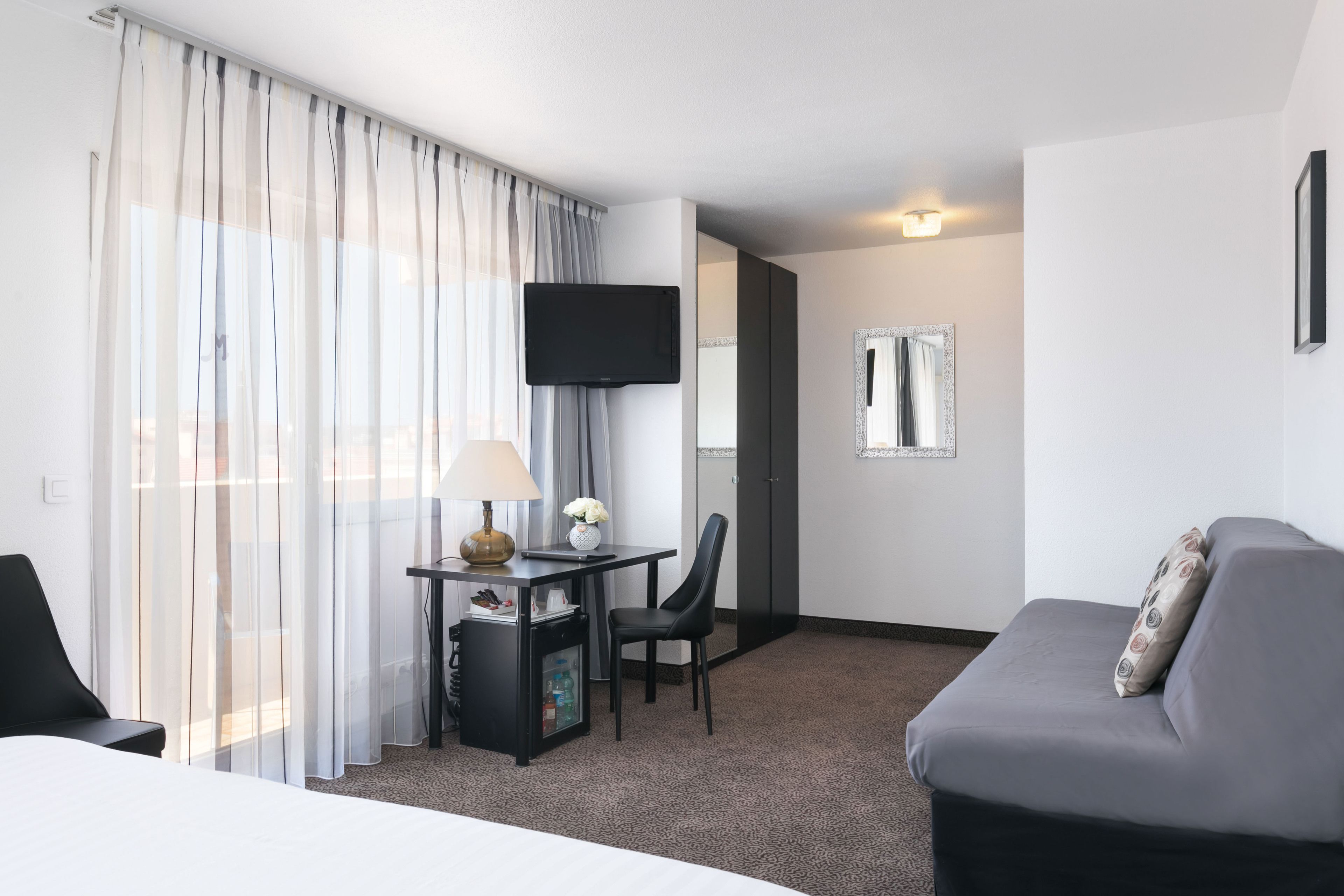 A hotel room with a bed, a desk with a lamp, a chair, a sofa, a TV, and a mirror.