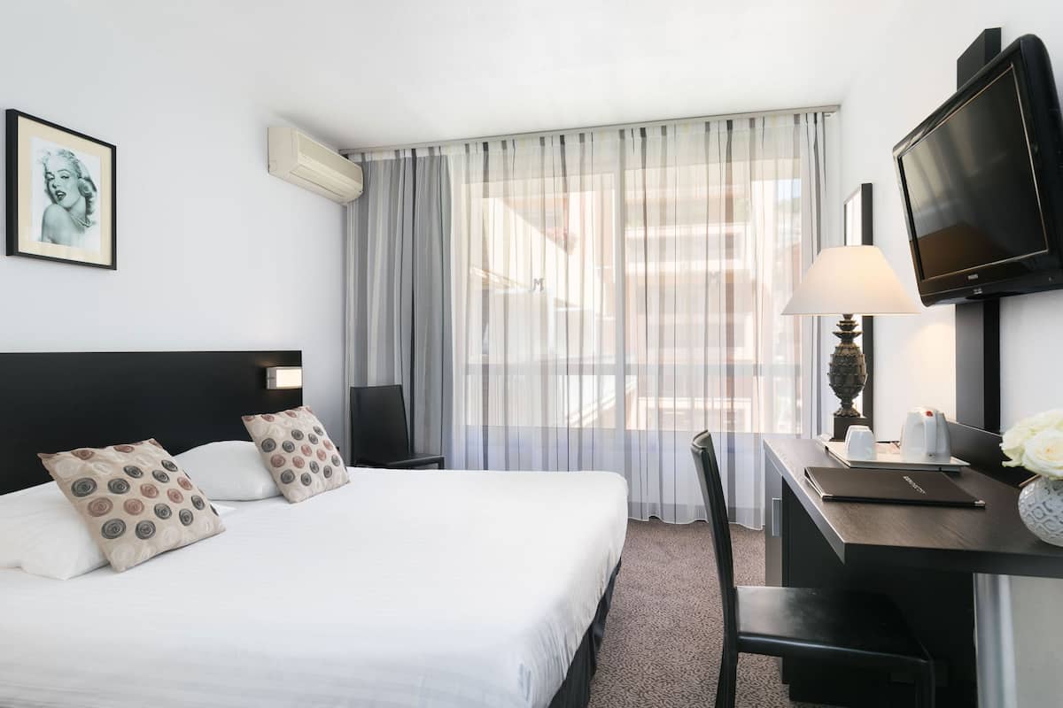 Ein Hotelzimmer mit Bett, Schreibtisch mit Lampe, Fernseher und Fenster mit durchsichtigen Vorhängen.