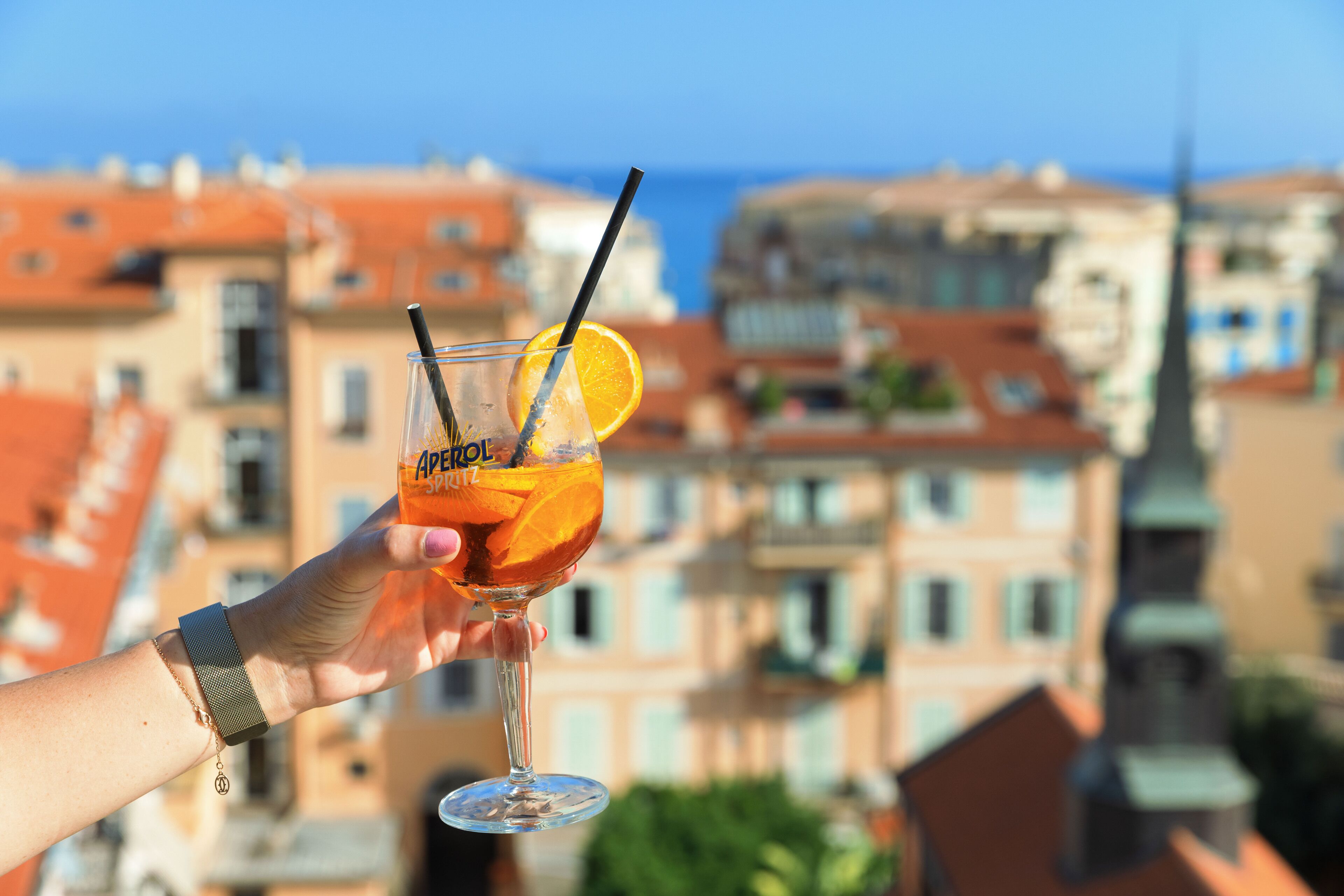 Una mano tiene un bicchiere da cocktail con una fetta d'arancia e una cannuccia, su uno sfondo di edifici con tetti rossi.