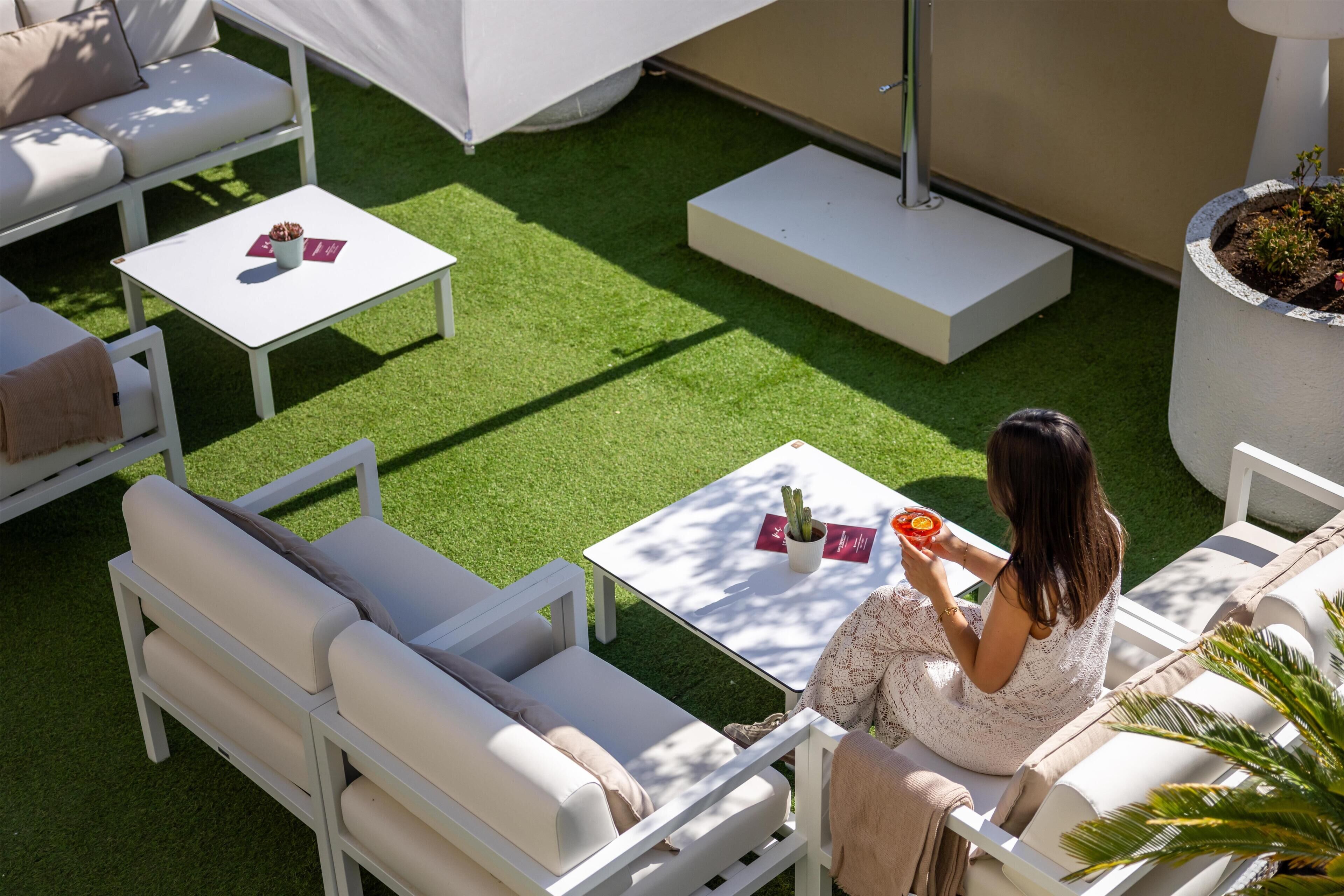 Een terras met witte meubels, waaronder een bank en twee tafels, en een vrouw die op een stoel zit.