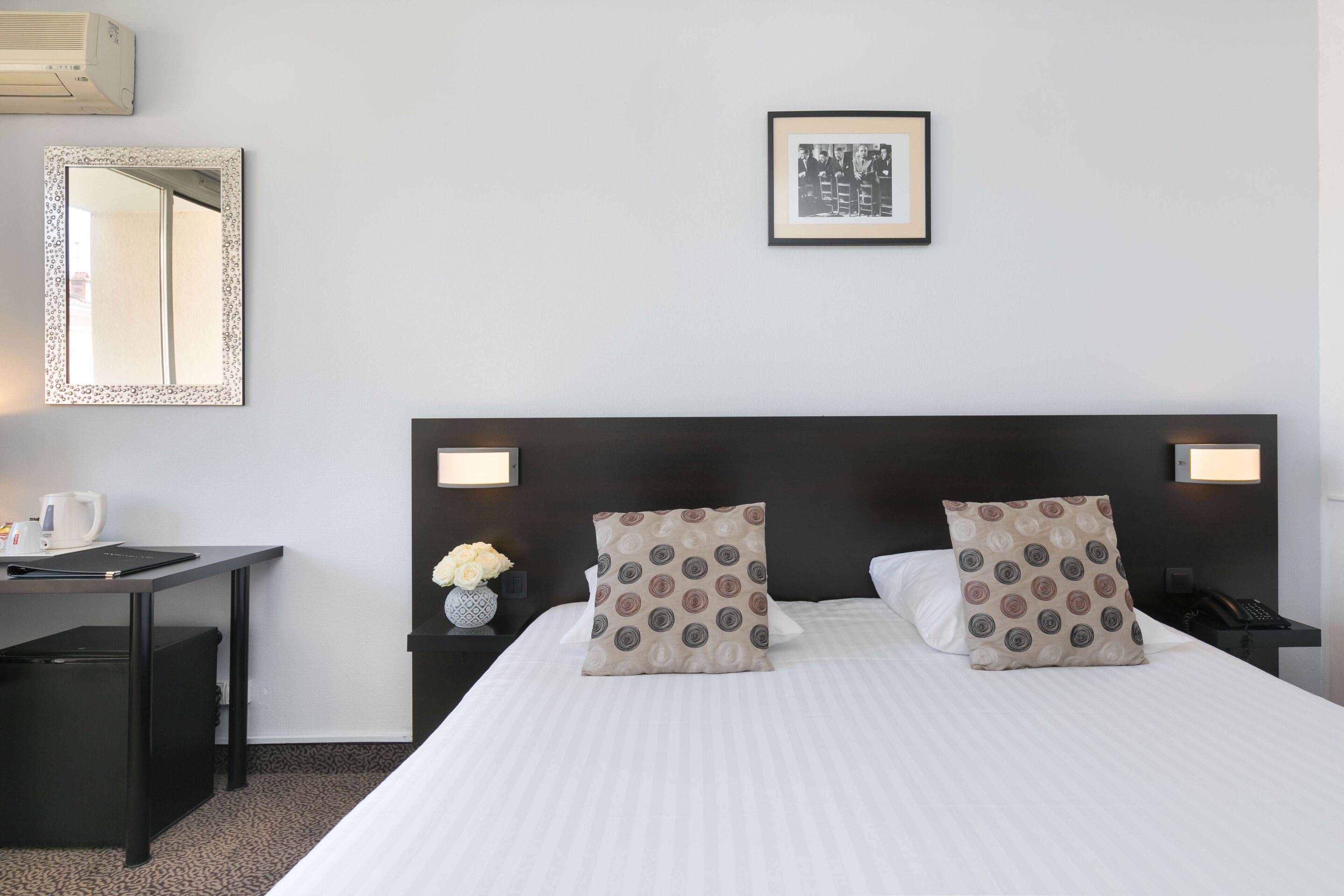 A hotel room with a bed, a desk, a chair, a nightstand, a mirror, and a framed picture on the wall.