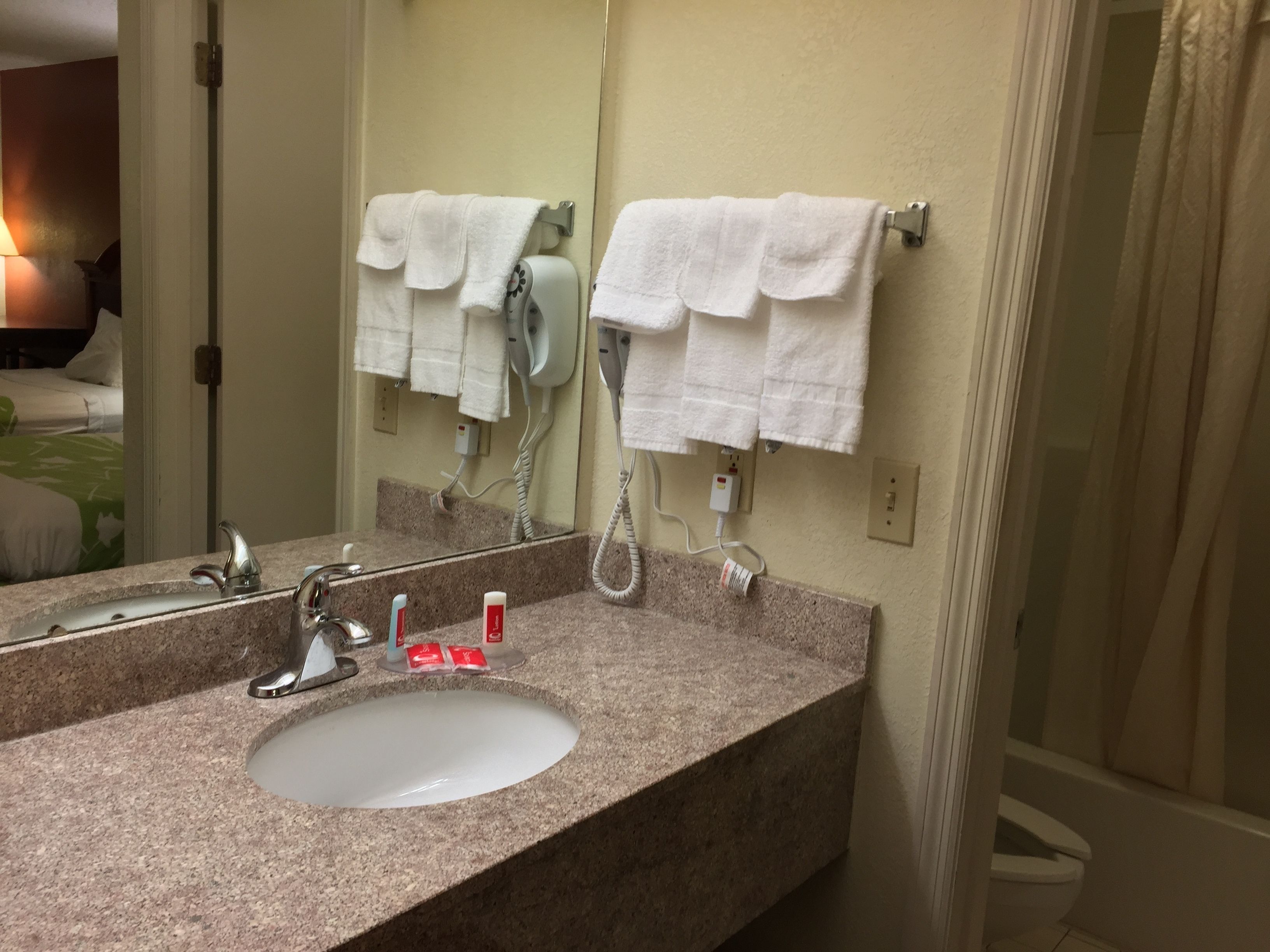 Un baño de hotel con lavamanos de encimera de granito, un espejo, un secador de pelo y un perchero con dos toallas blancas.