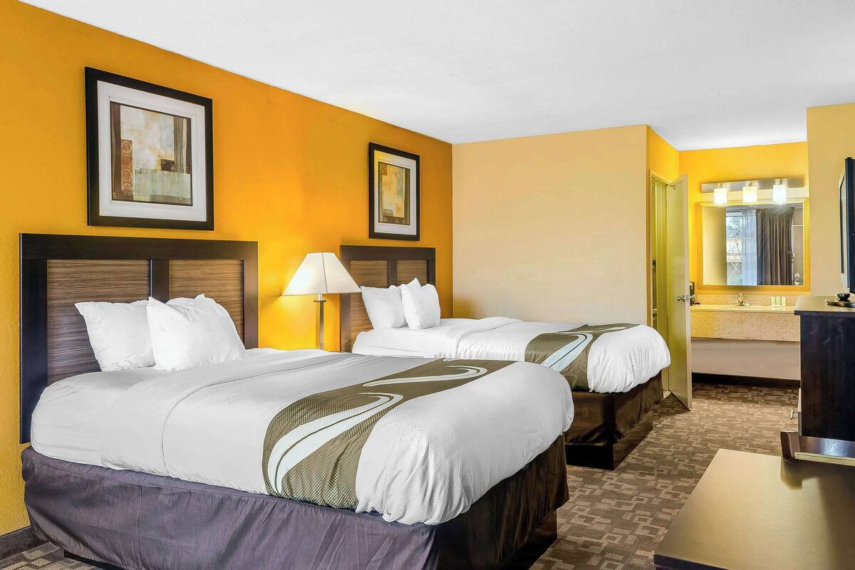 A hotel room with two beds, a bathroom with a mirror and sink, and artwork on the walls.