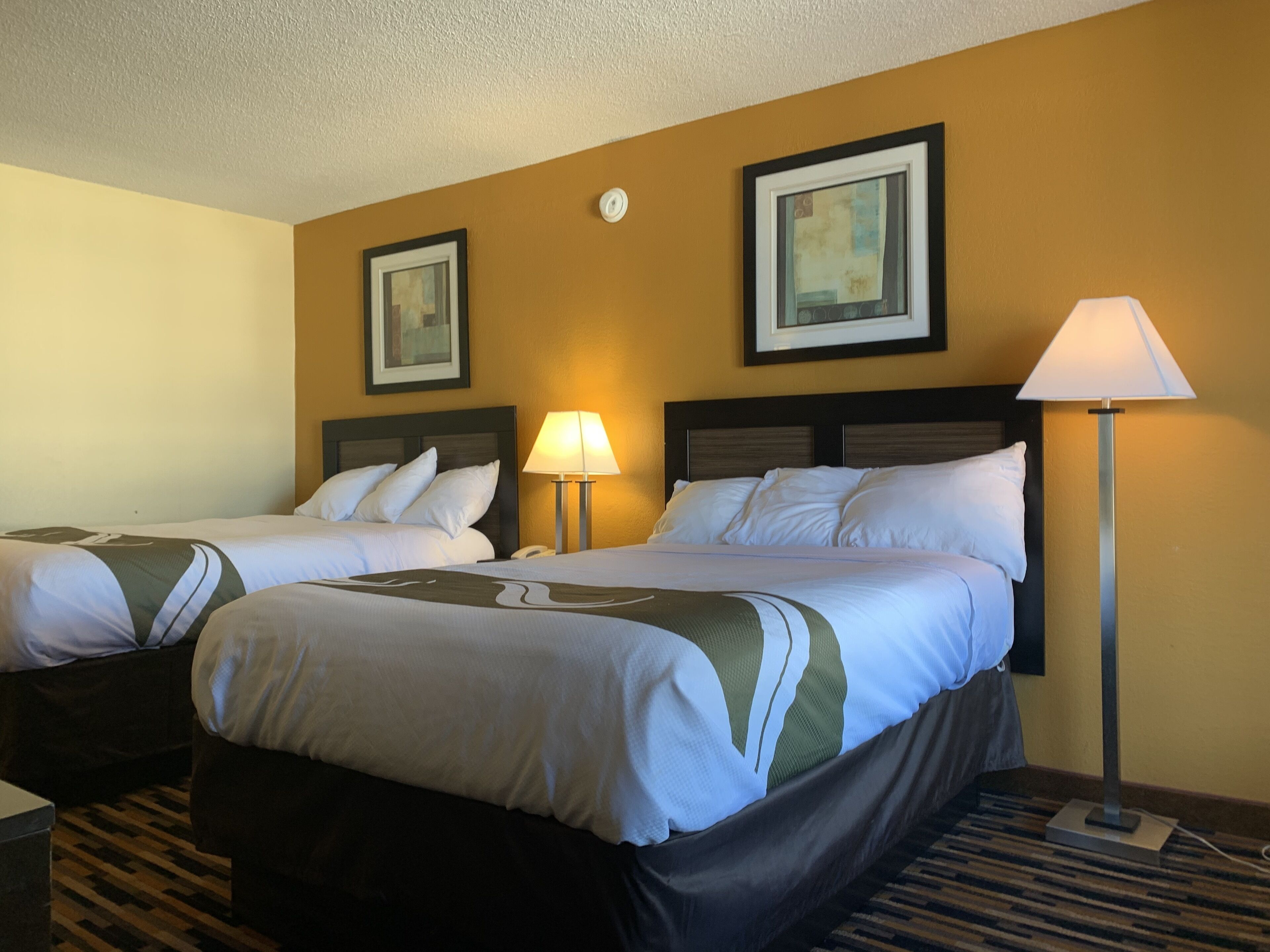 A hotel room with two beds, a nightstand, a lamp, and two framed pictures on the wall.