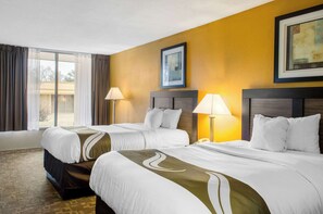 A hotel room with two beds, a large window with curtains, a framed picture on the wall, and a lamp beside each bed.