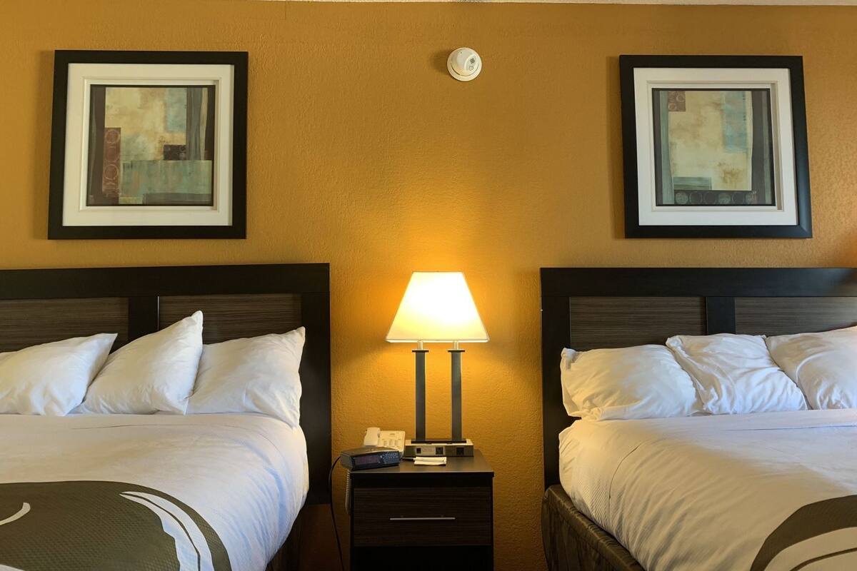 A hotel room with two beds, a nightstand with a lamp, and two framed artworks on the wall.