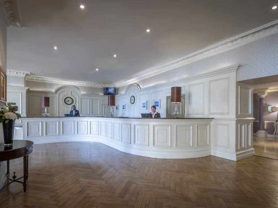 A reception area with a curved white reception desk, two receptionists, a clock, and a television on the wall.