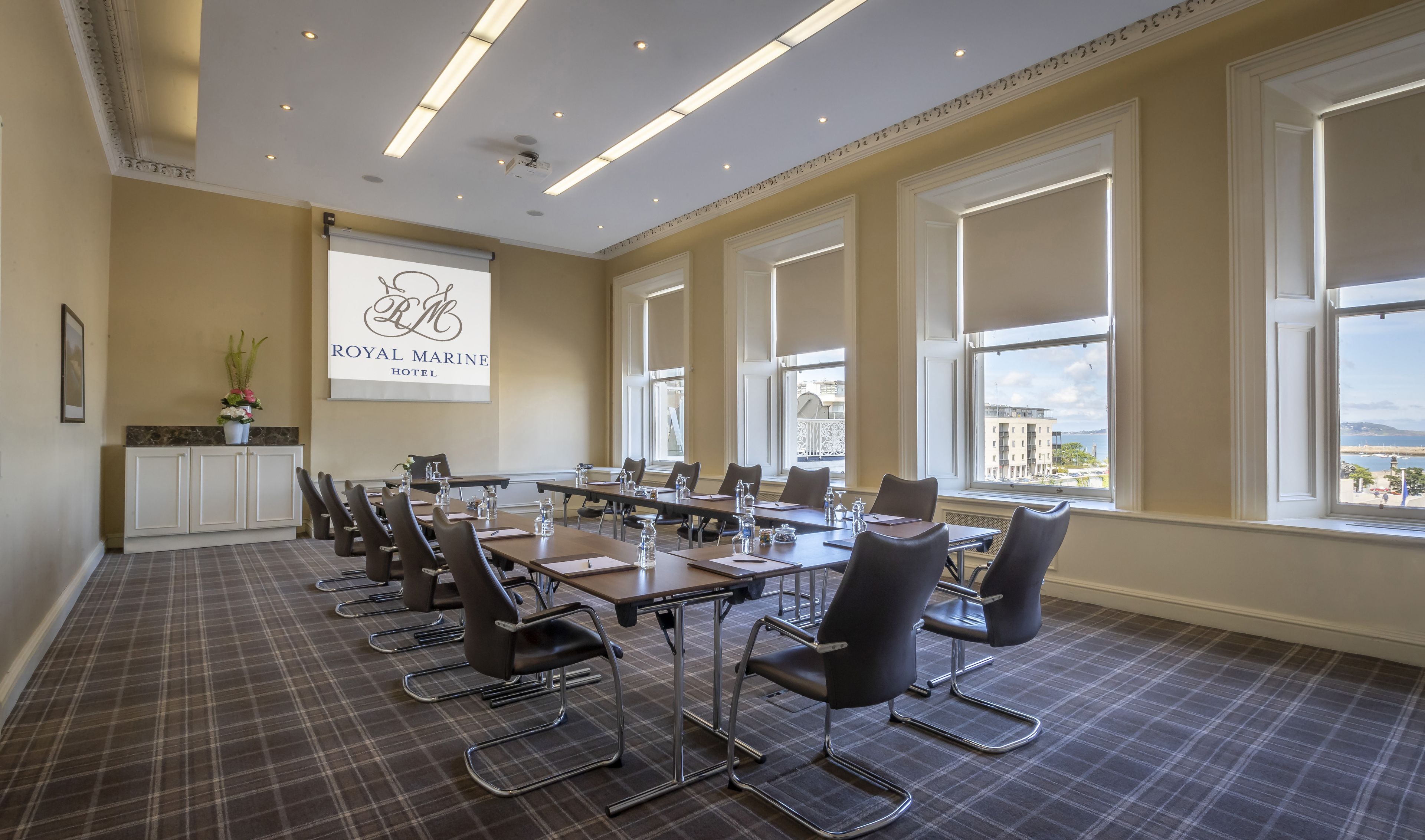 A conference room with a large table, chairs, a projector screen, and large windows with a view of buildings and the sea.