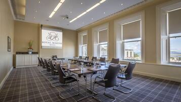 A conference room with a large table, chairs, a projector screen, and large windows with a view of buildings and the sea.