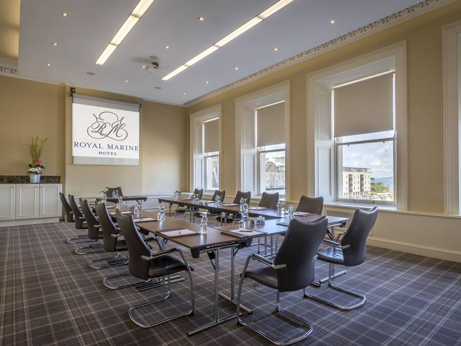 A conference room with a large table, chairs, a projector screen, and large windows with a view of buildings and the sea.