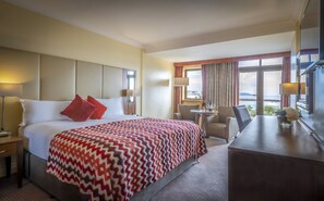 A hotel room with a bed, a desk, a chair, a lamp, and a view of the sea. - The Royal Marine Hotel (Dublin)