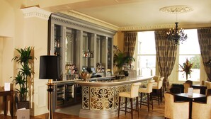 A bar area with a curved counter, bar stools, and a dining table with chairs. There are potted plants and a chandelier. - The Royal Marine Hotel (Dublin)